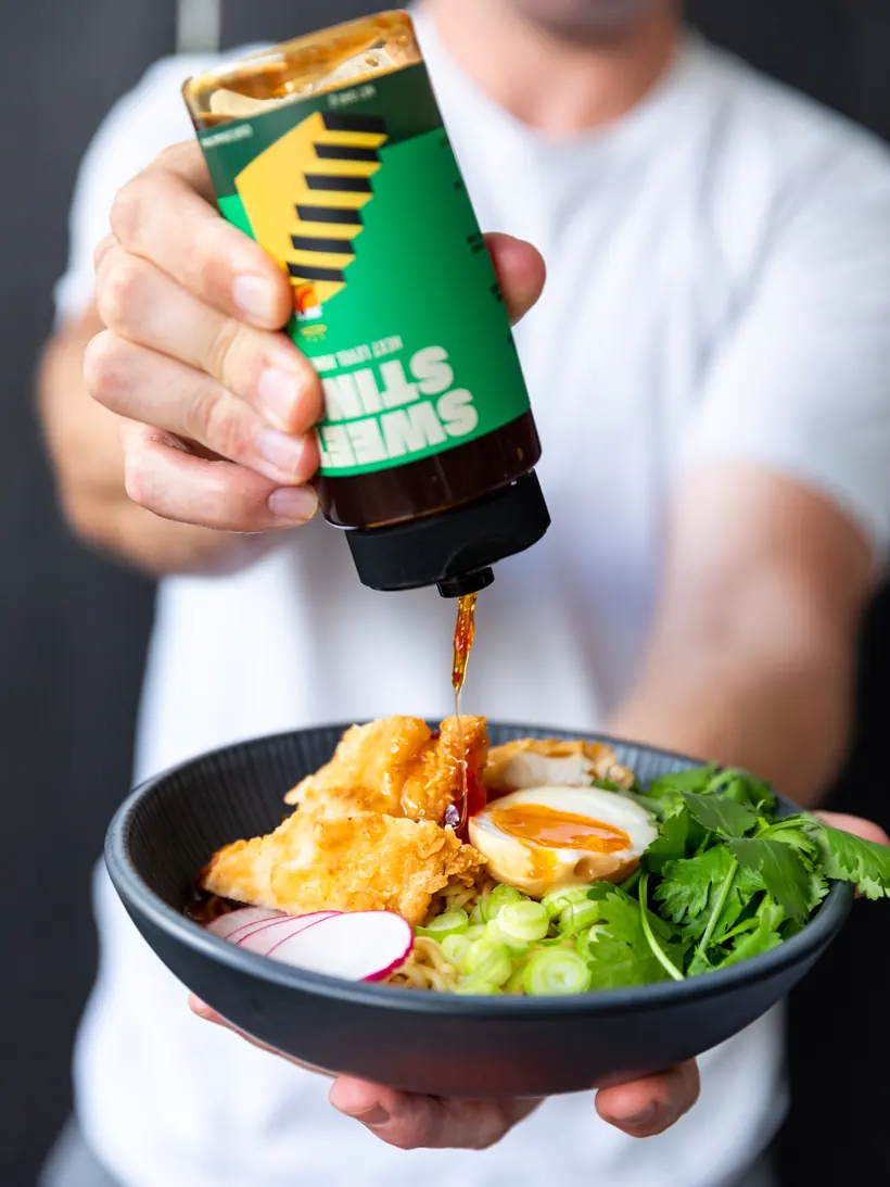 A bottle of Sweet Sting Hot Honey drizzled over a bowl of fried chicken ramen.